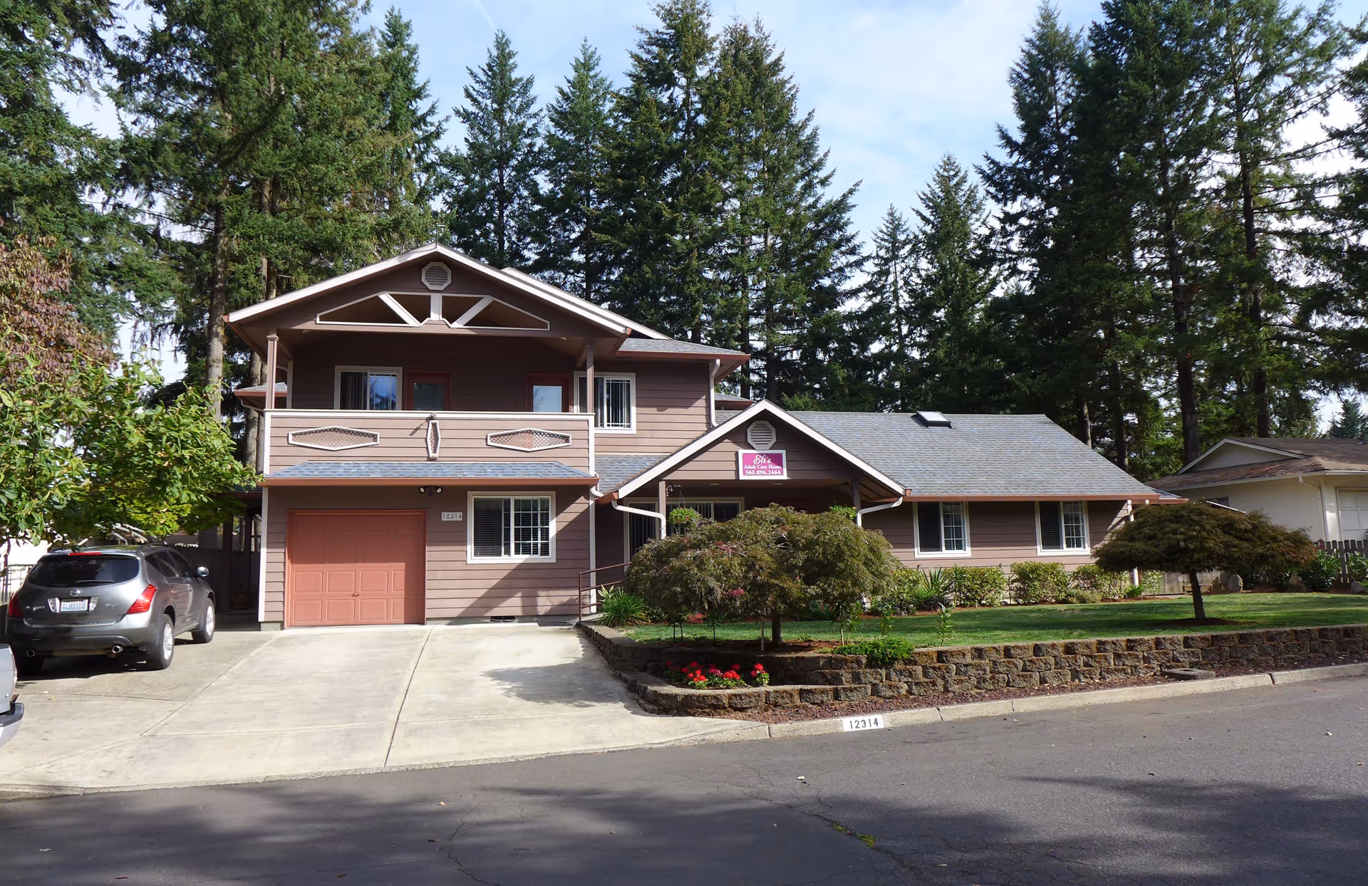 Exterior view of a two-story brown residential building with a garage, driveway, and well-maintained front yard with small trees and flowers. The building has a sign that reads 'Eli's Adult Care Home' above the entrance. Tall evergreen trees are visible in the background under a partly cloudy sky.