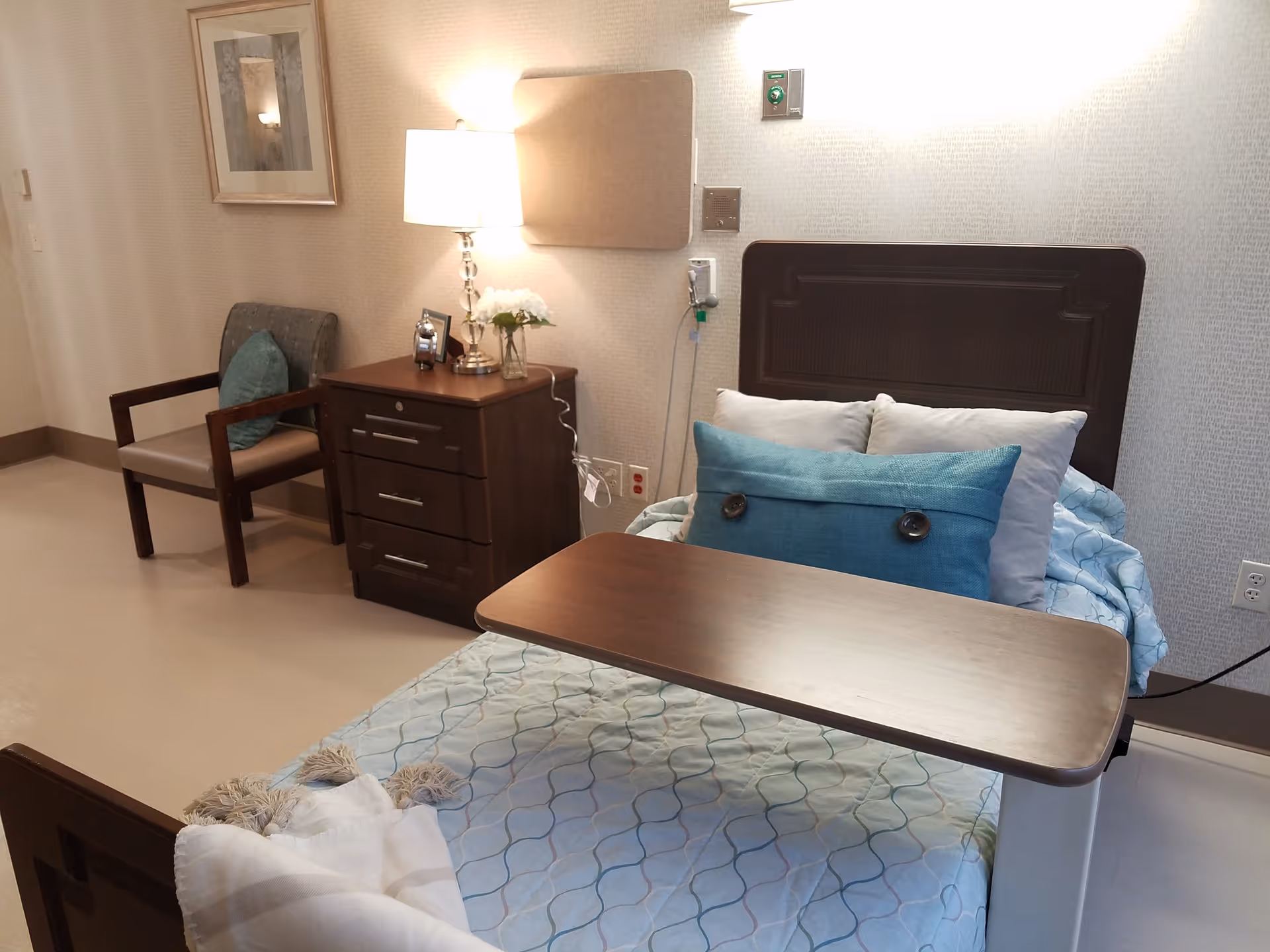 A neatly arranged senior living bedroom featuring a hospital-style bed with a blue patterned blanket and pillows, a wooden overbed table, a wooden nightstand with a lamp, flowers, and a clock, and a cushioned chair with a blue pillow. The room has light-colored walls with a framed picture and medical equipment mounted on the wall.
