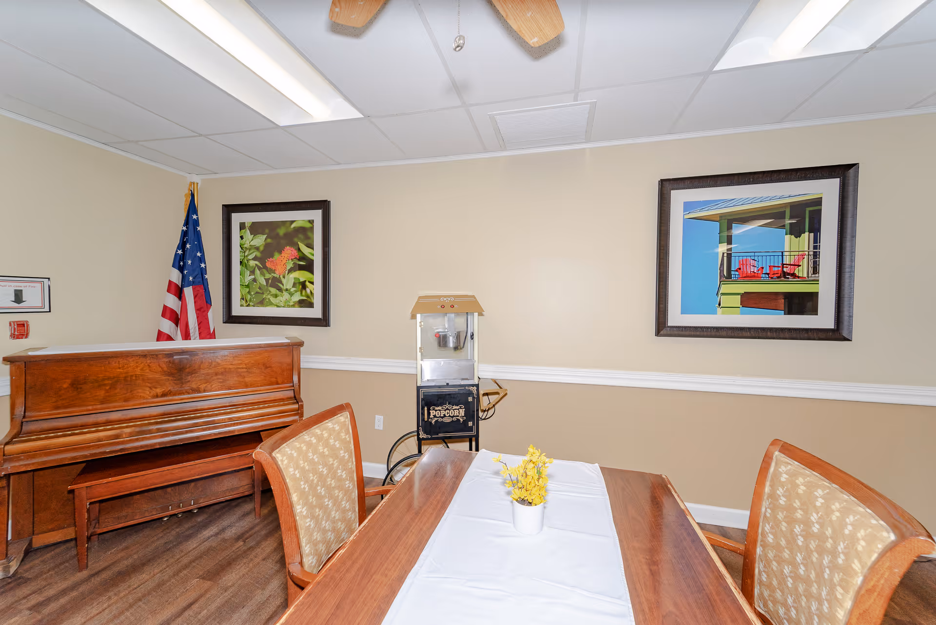 Dining room scene with table, chairs, piano, flag, and popcorn machine against beige walls.