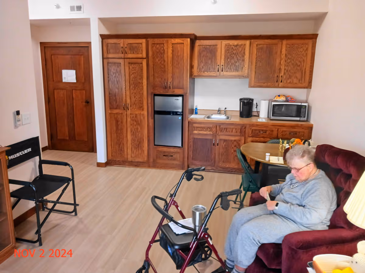 Senior resident seated in a burgundy chair beside a kitchenette with wooden cabinets and walker.
