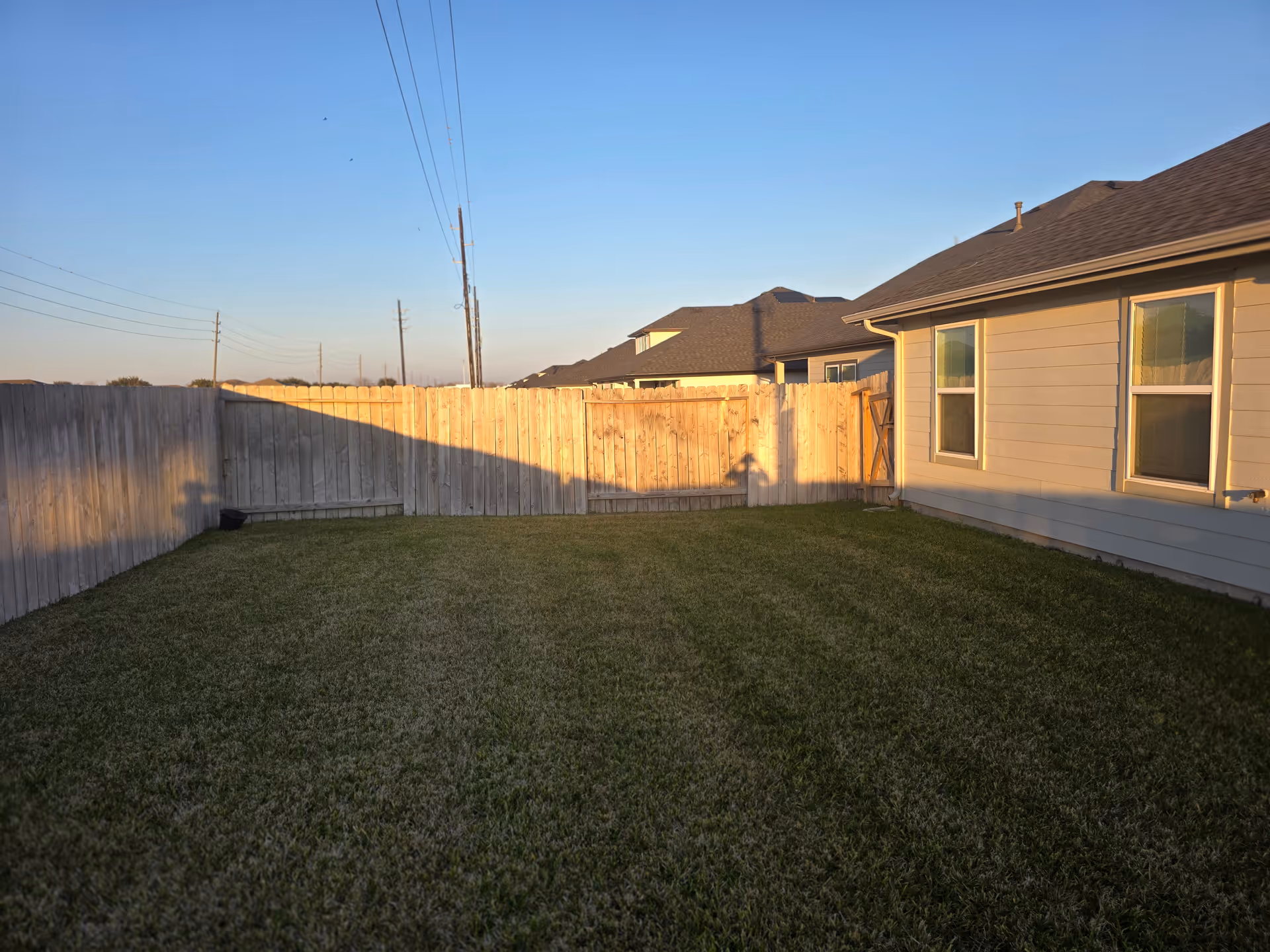 Sunlit backyard with a wooden fence, green lawn, and beige house with windows.