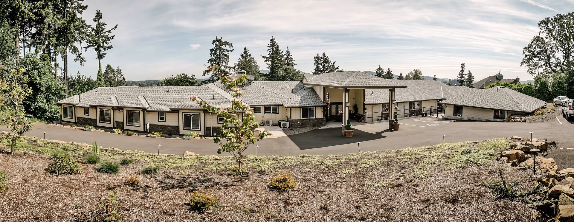 Exterior view of a single-story senior living facility building with a covered entrance, surrounded by trees and landscaped grounds under a partly cloudy sky.