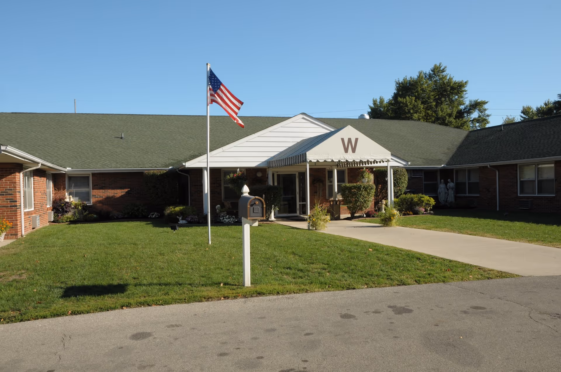 Front exterior view of a single-story brick building with a green roof and a white entrance canopy marked with a large letter 'W'. An American flag flies on a flagpole in the grassy front yard, and a mailbox is positioned near the driveway leading to the entrance. Trees and shrubs surround the building under a clear blue sky.
