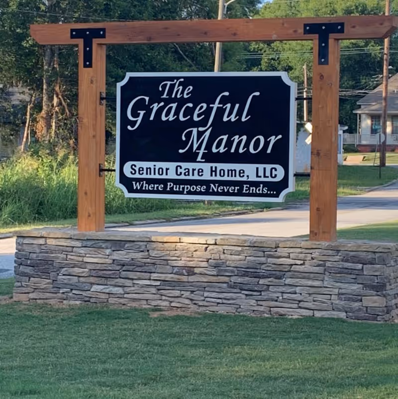 A large outdoor sign for The Graceful Manor Senior Care Home, LLC, mounted on a wooden frame with a stone base, located near a road with grass and trees in the background.