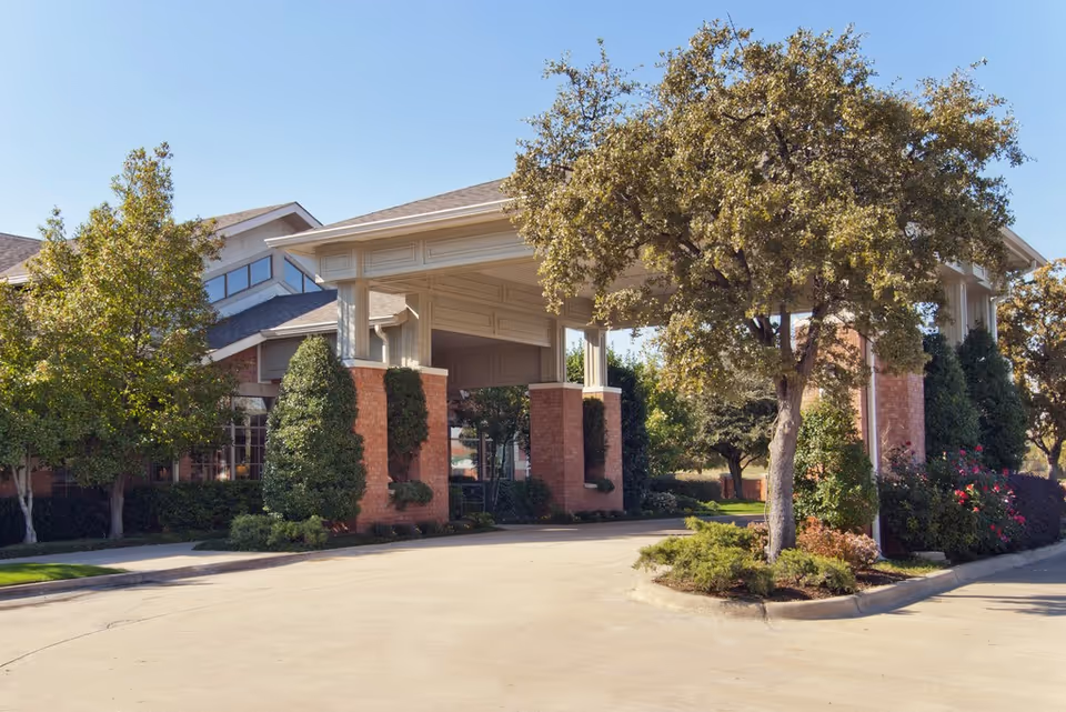 Exterior view of The Remington at Valley Ranch facility entrance with a covered driveway, surrounded by trees and shrubs under a clear blue sky.