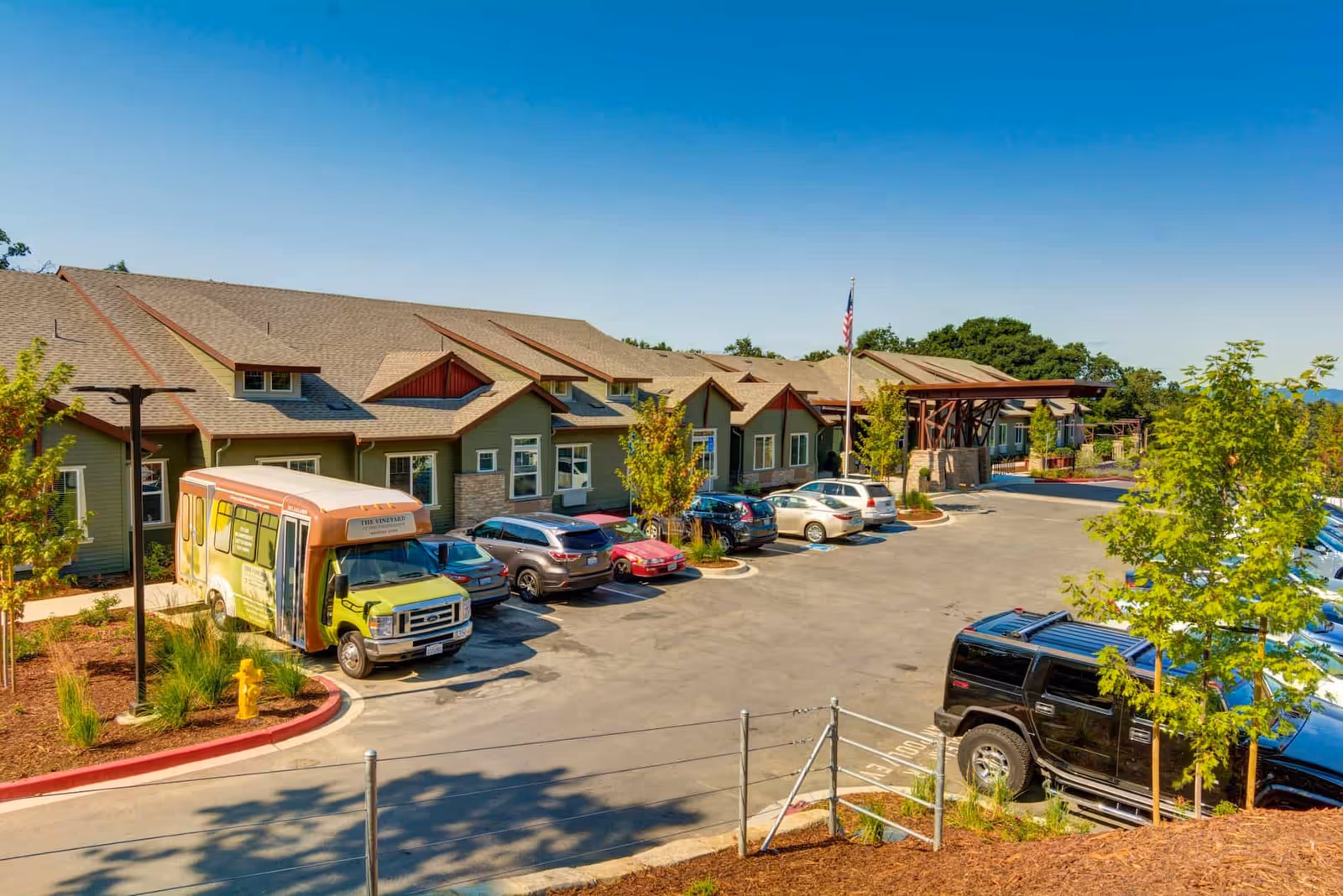 A single-story memory care facility building with a parking lot, several cars and a shuttle bus, landscaped grounds, and a clear blue sky.