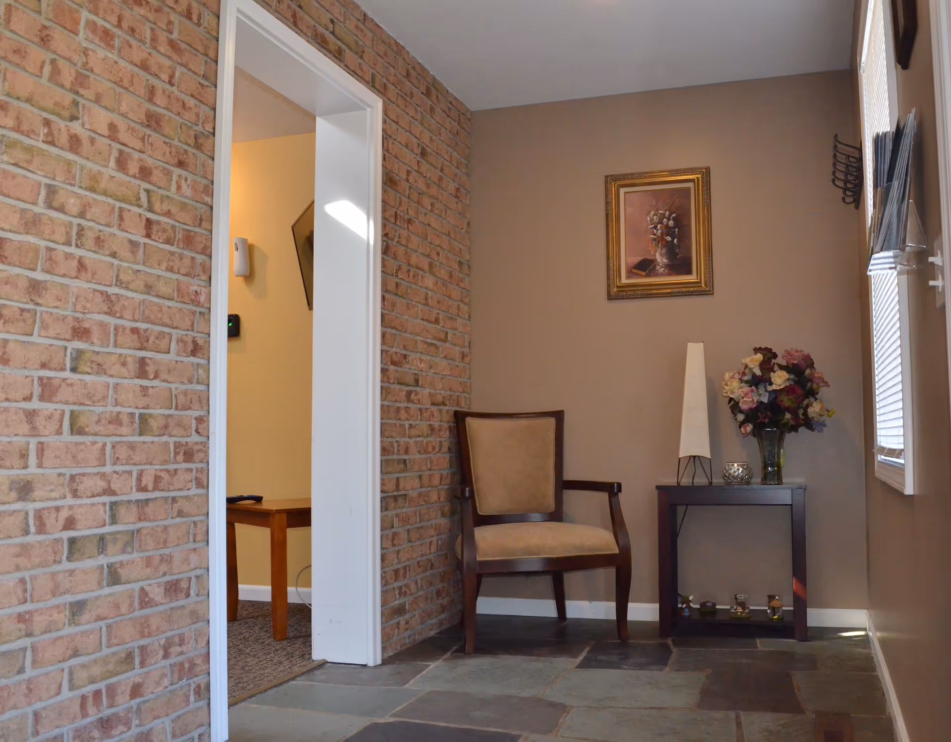 A small foyer with brick wall, chair, side table, flowers, and wall art.