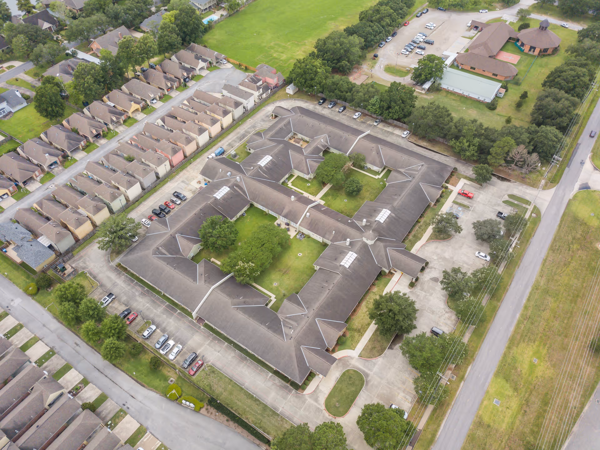 Aerial view of a senior living facility named Brookdale Lafayette, showing a large building with a central courtyard surrounded by parking lots and nearby residential houses and green fields.