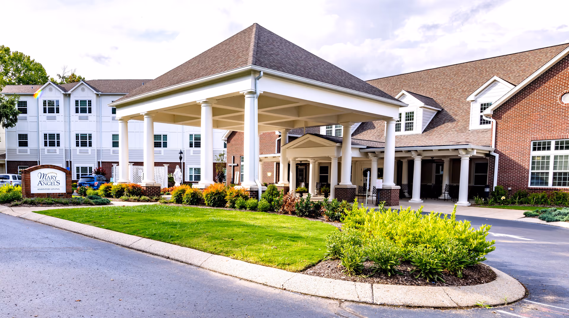 Exterior view of Mary Queen of Angels Assisted living facility showing a large covered entrance with white columns, well-maintained green lawn, shrubs, and a sign with the facility's name. The building has a combination of brick and white siding with multiple windows and a sloped roof.