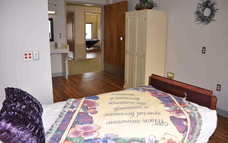 Bedroom in a care facility with bed, floral blanket, dresser, and open doorway.