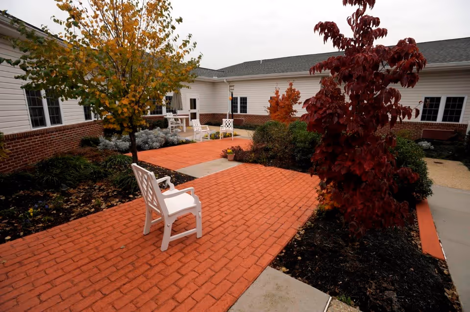Outdoor courtyard area with red brick pathways, white chairs, and various trees with autumn foliage surrounded by a single-story building with white siding and brick accents.