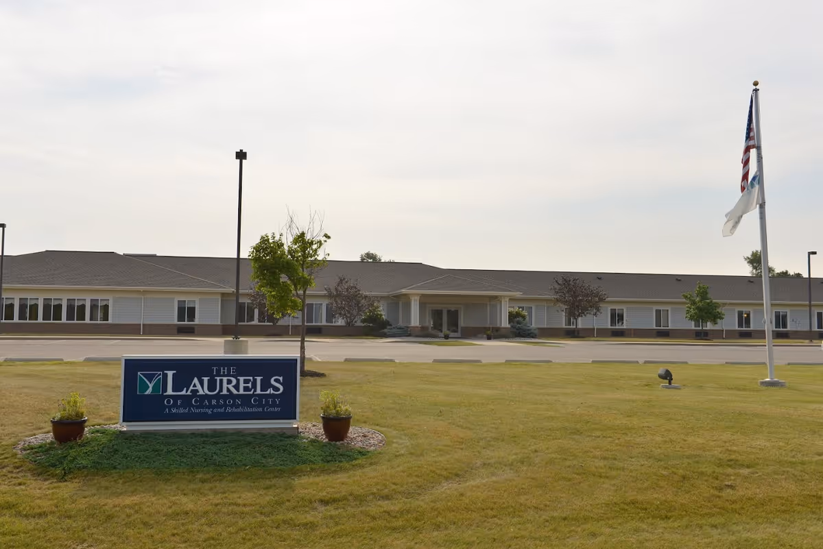 Exterior view of The Laurels of Carson City, a single-story building with a large lawn in front. There is a sign on the lawn that reads 'The Laurels of Carson City, A Skilled Nursing and Rehabilitation Center' and an American flag on a flagpole to the right.