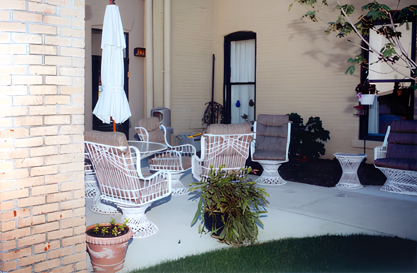 Outdoor patio area with white metal chairs and tables, some with cushions, a closed white umbrella, potted plants, and a beige brick wall and building exterior in the background.