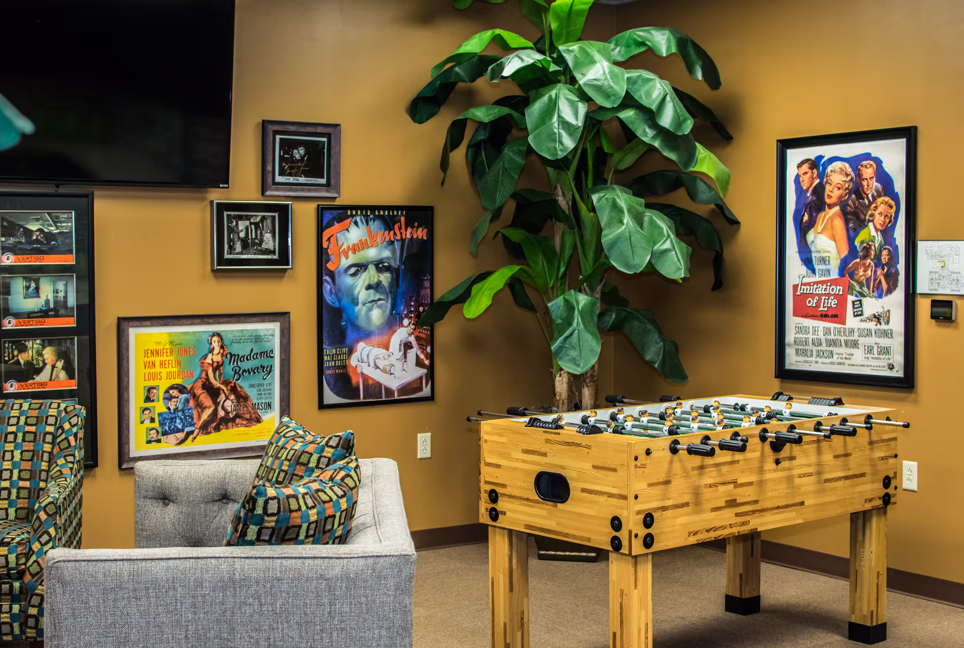 Warm yellow lounge with a foosball table, plant, framed movie posters, and gray sofa.