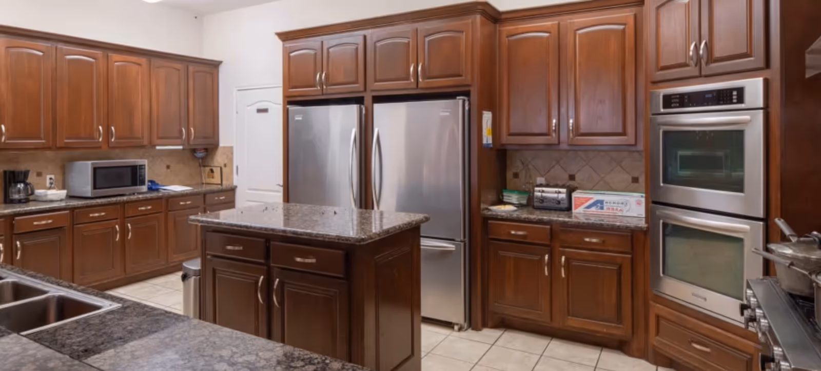 Spacious kitchen with dark wood cabinetry, a central island, stainless steel double-door refrigerator, and double wall ovens.