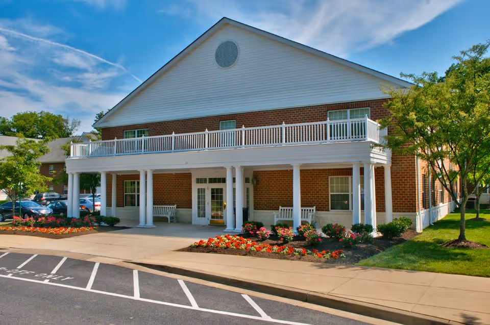 Two-story brick building with white columns, a covered entrance and balcony, and landscaped flower beds in front.