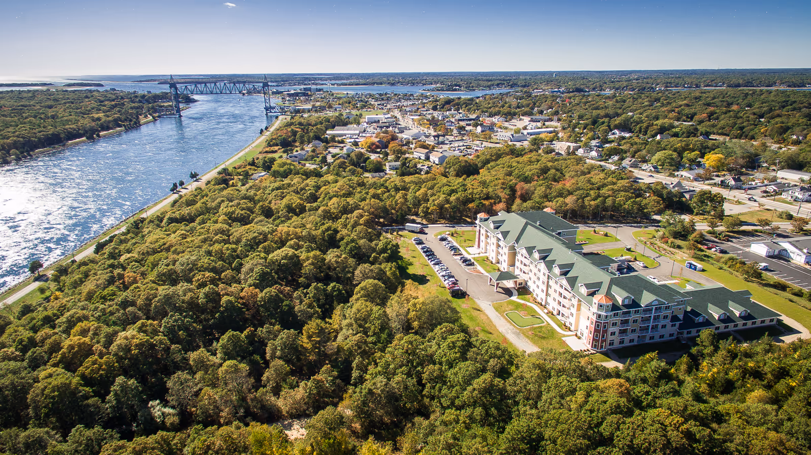 Aerial view of Keystone Place at Buzzards Bay, a large multi-story building surrounded by dense green trees, with a river and a bridge visible to the left and a town in the background under a clear blue sky.