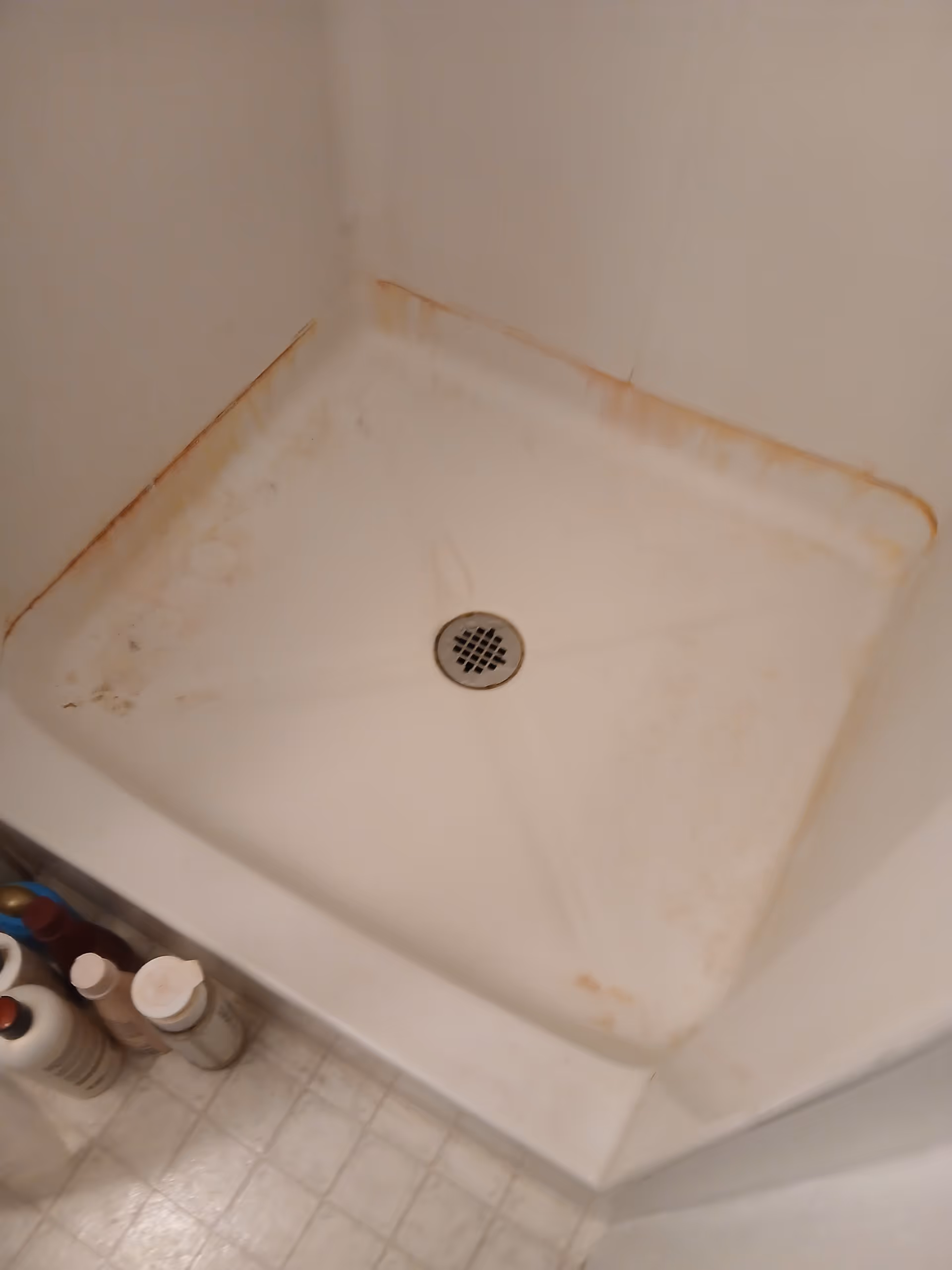 A white shower base with a drain, brown stains along edges, and cleaning bottles nearby.