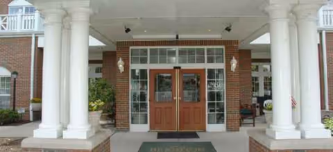 A brick senior living entrance with white columns and wooden double doors under a covered portico.