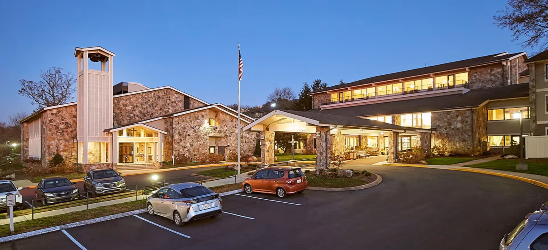 Front exterior of Alexian Village of Tennessee at dusk showing a stone-clad senior living building with a covered entrance, flagpole and parked cars.