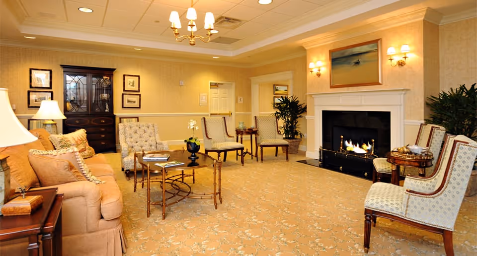 A cozy living room with beige walls and carpet, featuring a fireplace with a painting above it. The room is furnished with a tan sofa, several upholstered chairs, wooden side tables, a coffee table with books and a small plant, and decorative lamps. There are framed pictures on the walls and potted plants in the corners.