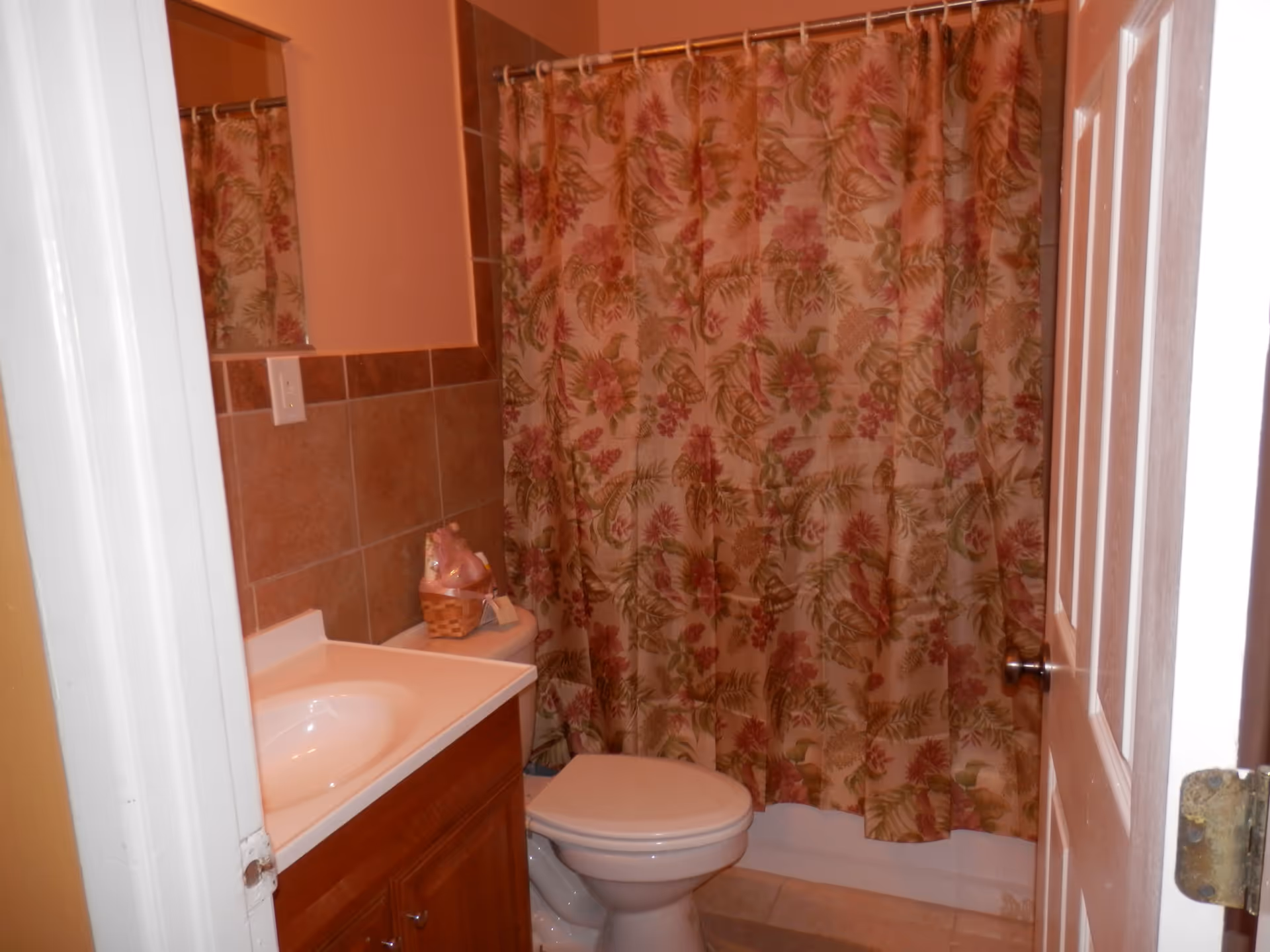 Small bathroom with a white toilet, wooden vanity with a white sink, and a shower with a floral patterned curtain in shades of pink, green, and beige. The walls are painted peach and partially tiled with brown tiles. A mirror is partially visible above the sink.