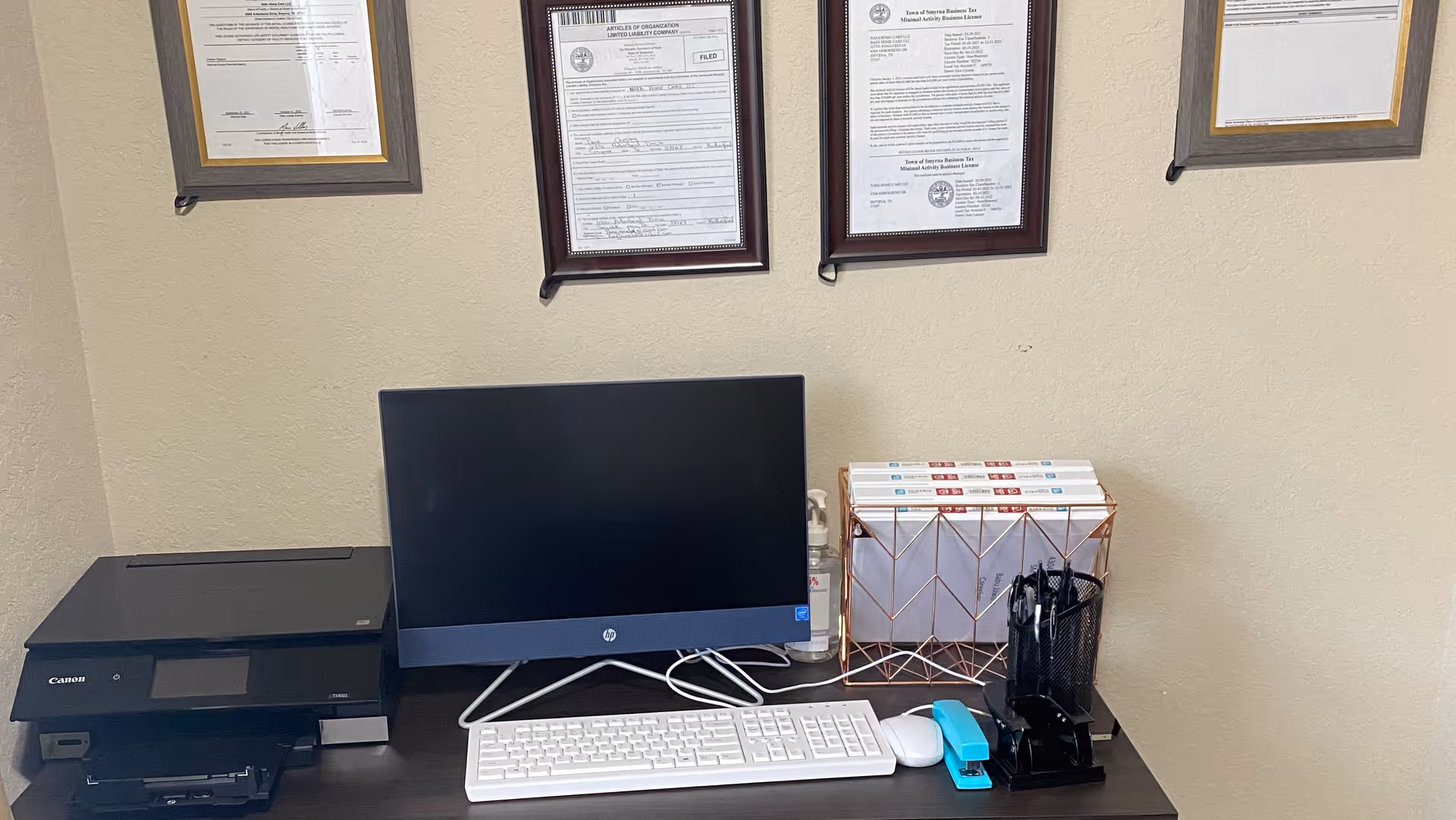 Office desk with computer monitor, keyboard, printer, and organized paperwork against framed certificates.