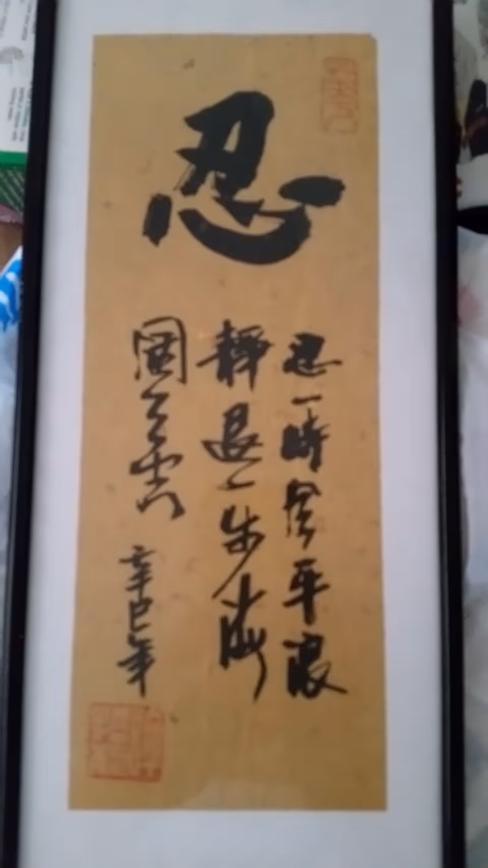 Framed vertical calligraphy scroll with black Chinese characters hanging indoors.