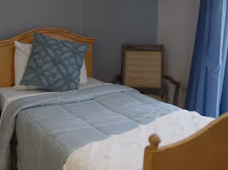 A neatly made bedroom with a wooden bed, blue bedding, curtains, and a chair.
