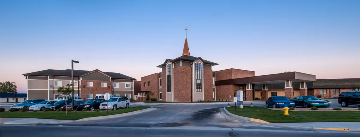 Front exterior of Ascension Living Via Christi Village with brick buildings and parked cars.