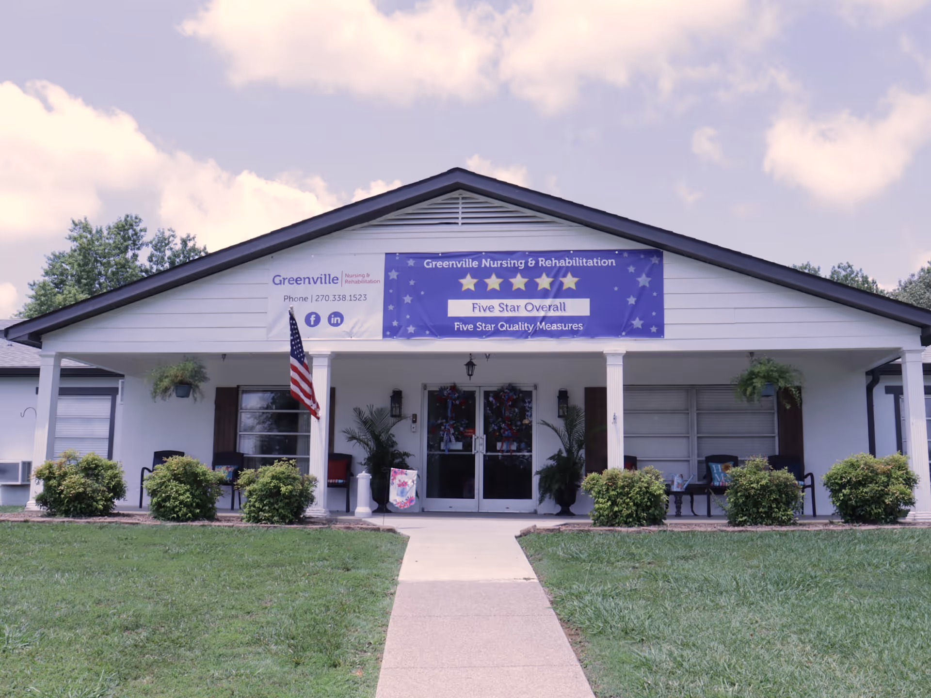 Front exterior view of Greenville Nursing and Rehabilitation building with a walkway leading to the entrance. The building has a white facade with a blue banner displaying five stars and the text 'Five Star Overall, Five Star Quality Measures'. There are bushes and an American flag near the entrance, along with some chairs and potted plants on the porch.