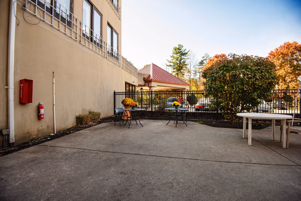 Outdoor patio area at Fairview Nursing and Rehabilitation Center with concrete flooring, metal tables and chairs, potted plants with flowers, a large bush, and a beige building wall with windows. There is a black metal fence separating the patio from a parking lot with cars and trees with autumn foliage in the background.