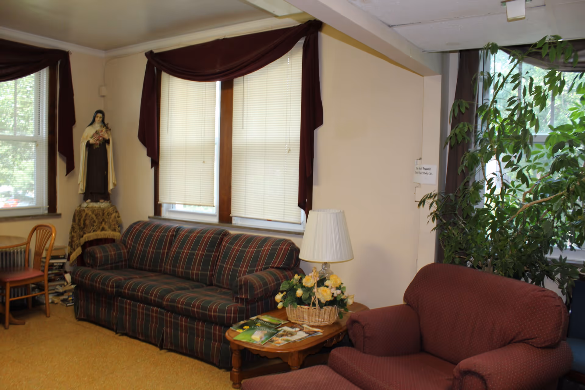A cozy living room area with a plaid sofa, a maroon armchair, a wooden side table with a lamp and a basket of yellow flowers, and a large green plant near the window. There is a statue of a religious figure on a small table covered with a decorative cloth in the corner. The windows have beige blinds and maroon valances.