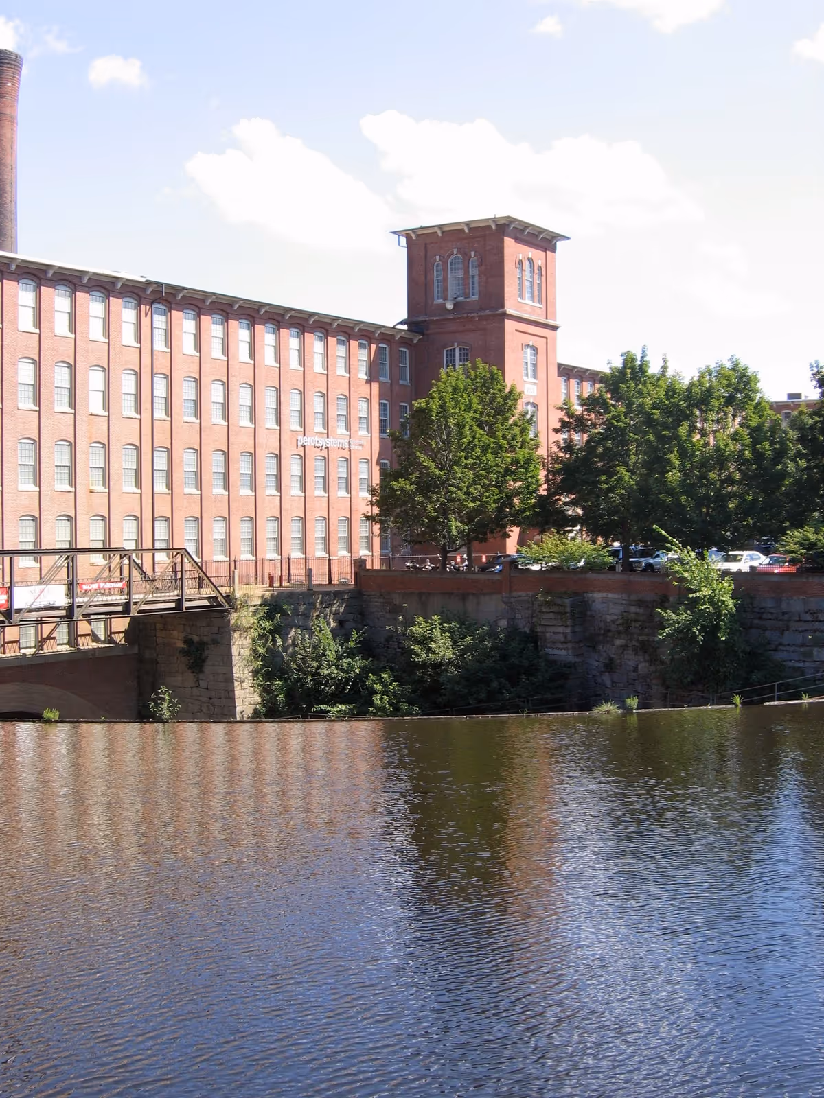 Brick multi-story building beside a river with trees and a small bridge.
