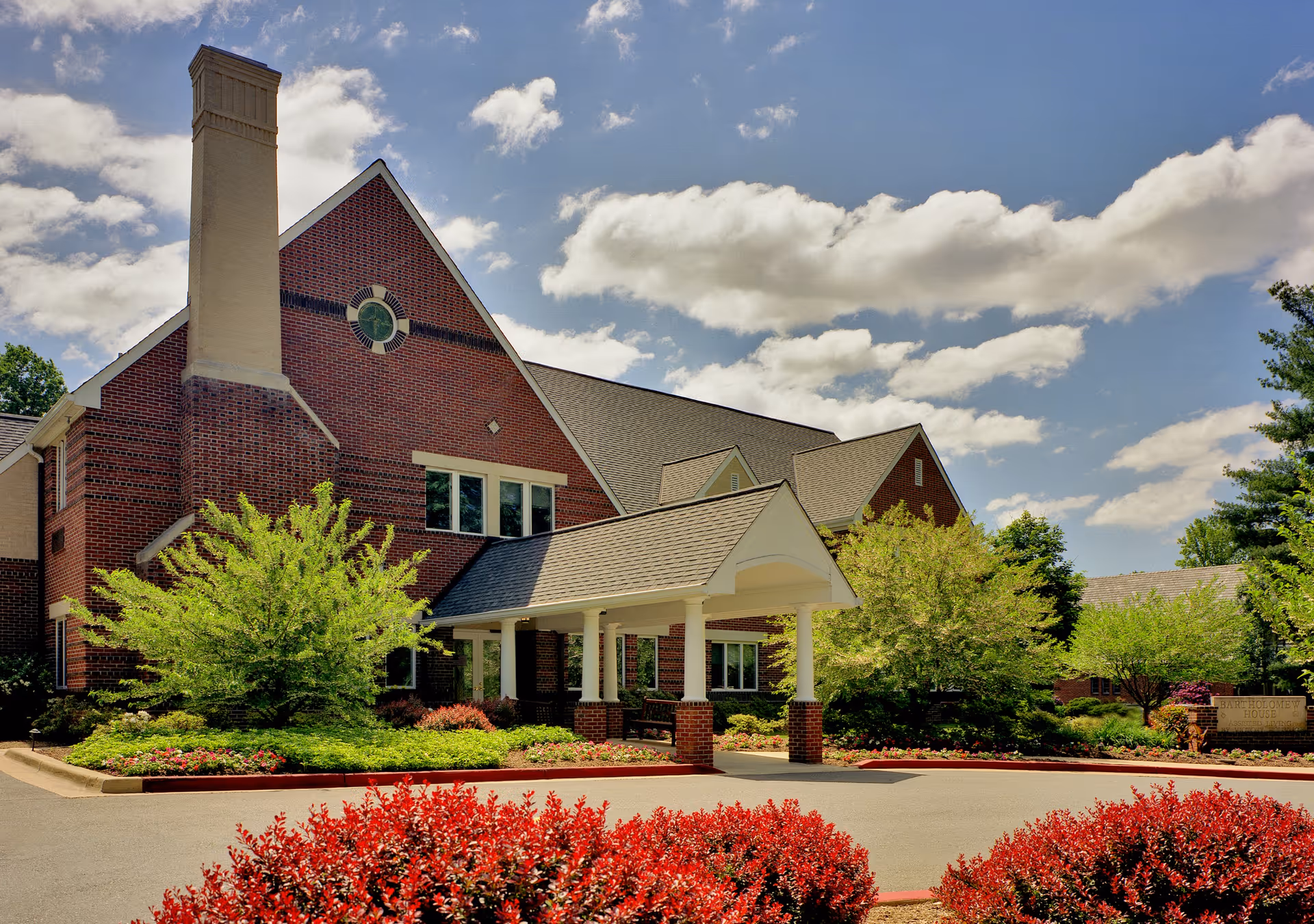 Brick senior living building front with a covered porte-cochère, landscaped beds, and a blue sky.