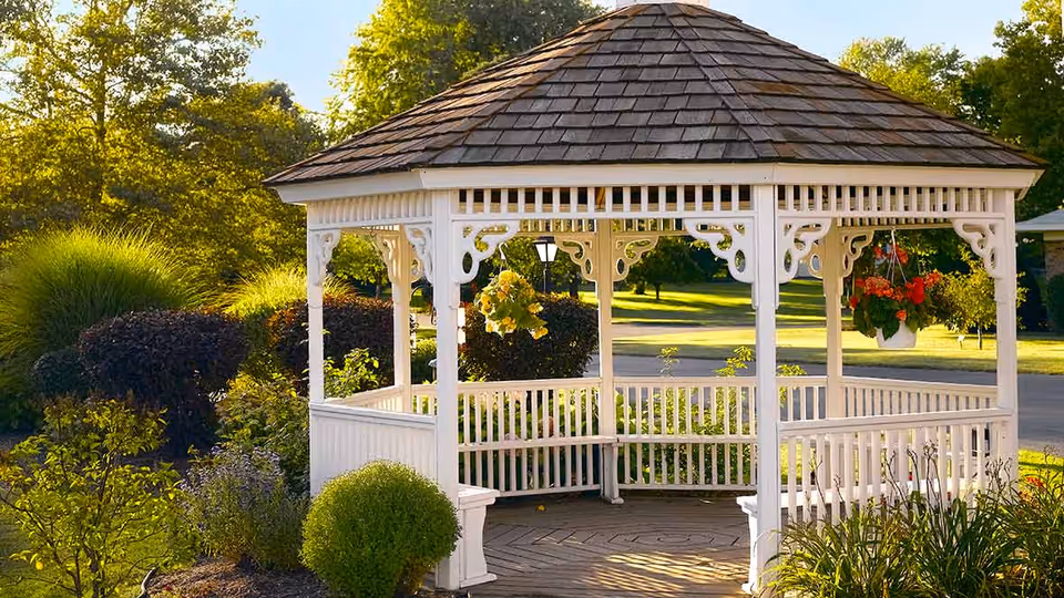 A white wooden gazebo with a shingled roof situated in a garden area with green bushes, hanging flower pots, and trees in the background under a sunny sky.