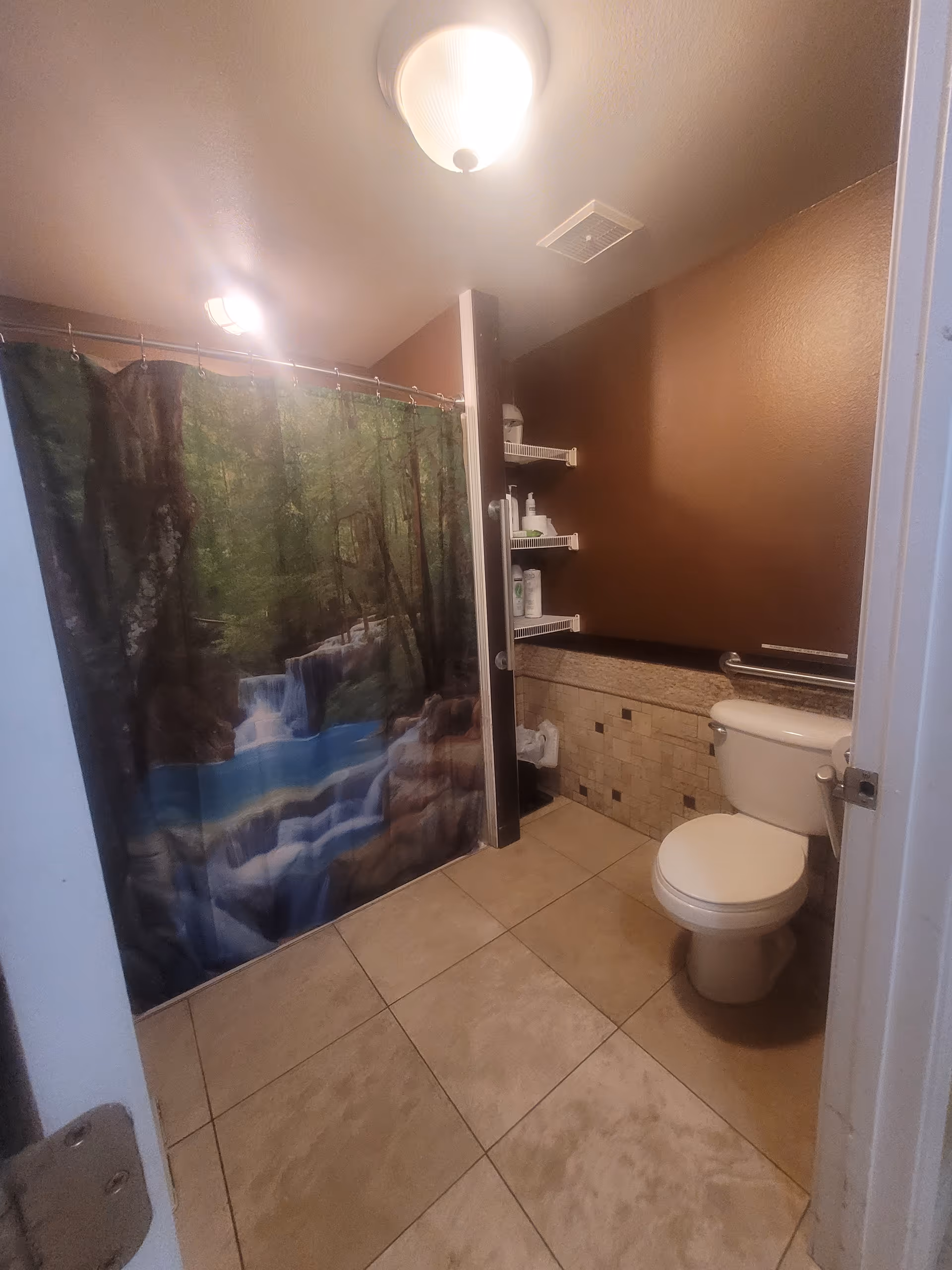Bathroom with forest themed shower curtain, toilet, tiled floor, and wall shelves.