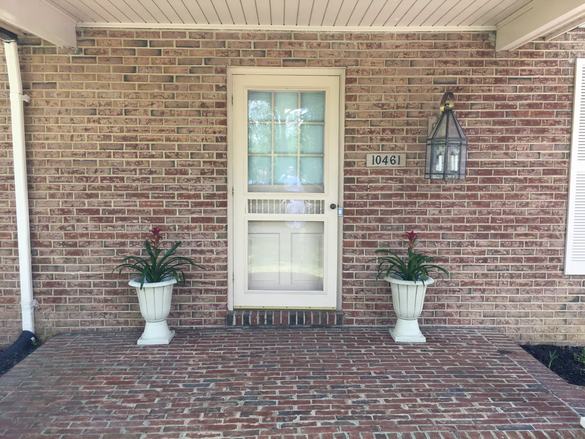 Front entrance of a brick building with a white door featuring glass panels. Two white planters with green plants and red flowers are placed symmetrically on either side of the door. A lantern-style light fixture is mounted on the wall to the right of the door, and the number 10461 is displayed on a plaque next to the light.