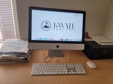 iMac computer on a wooden desk displaying KWATE HOME CARE logo in an office.
