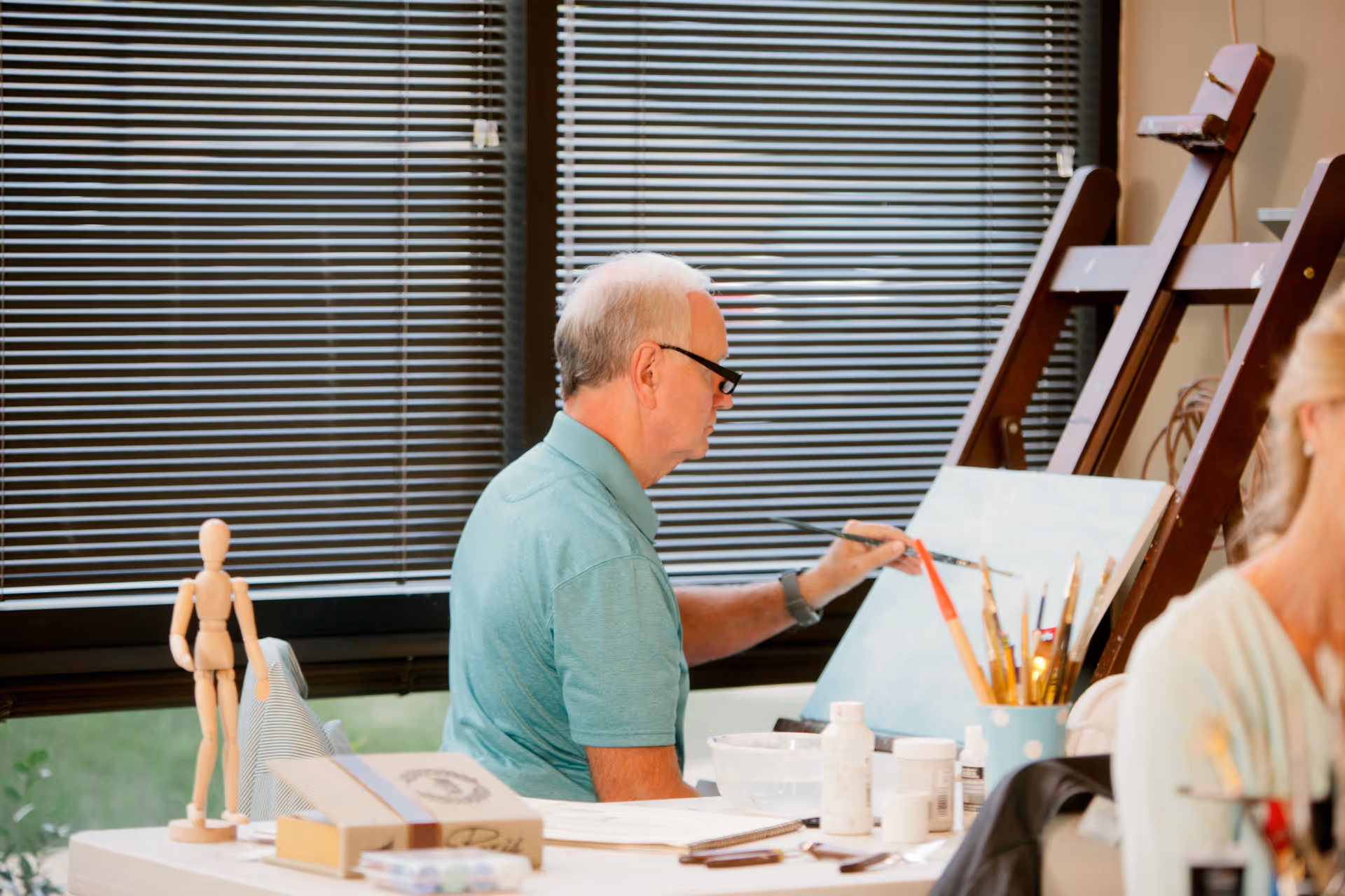 An elderly man wearing glasses and a teal shirt is painting on a canvas in a well-lit room with large windows covered by blinds. Various art supplies, including paintbrushes, paint bottles, and a wooden mannequin, are on the table in front of him. Another person is partially visible on the right side of the image.