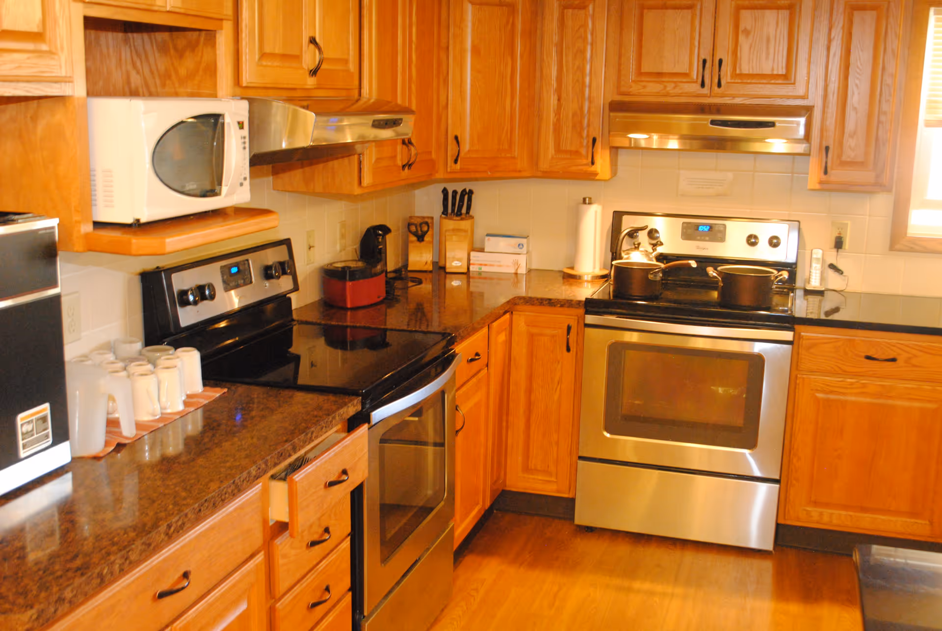 A kitchen with wooden cabinets, a microwave, two stainless steel electric stoves with pots on them, a coffee maker, a knife block, paper towels, and several white mugs and pitchers on the countertop.