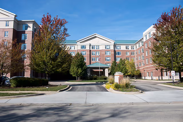 Brick senior living building with a green roof, entrance canopy, and autumn trees.