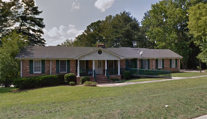 Single-story brick senior living facility with front porch and steps, surrounded by lawn.