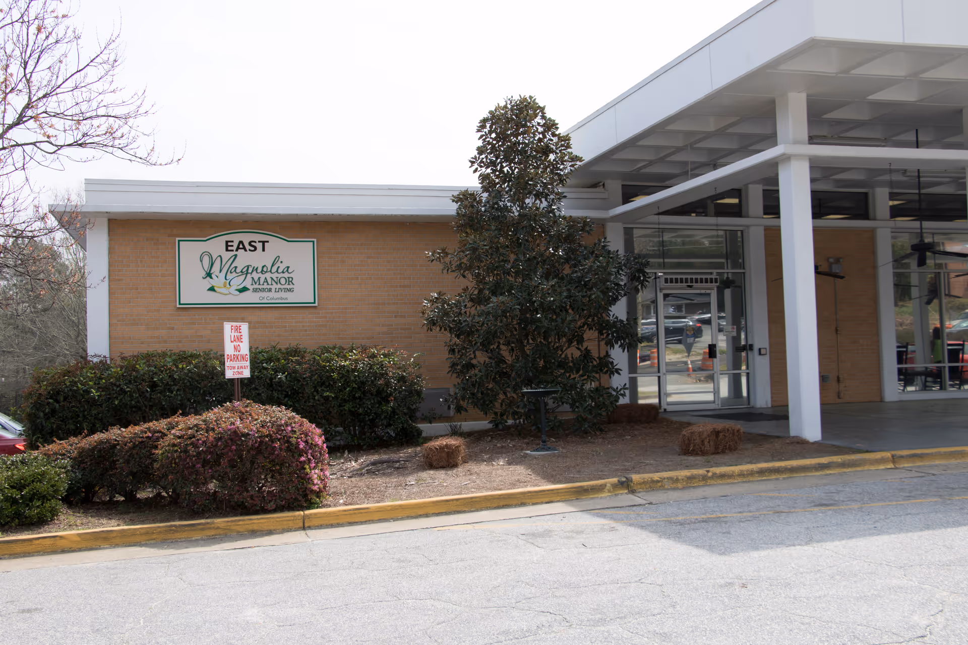 Exterior view of Magnolia Manor nursing center entrance with sign, brick wall, glass doors, and landscaping.