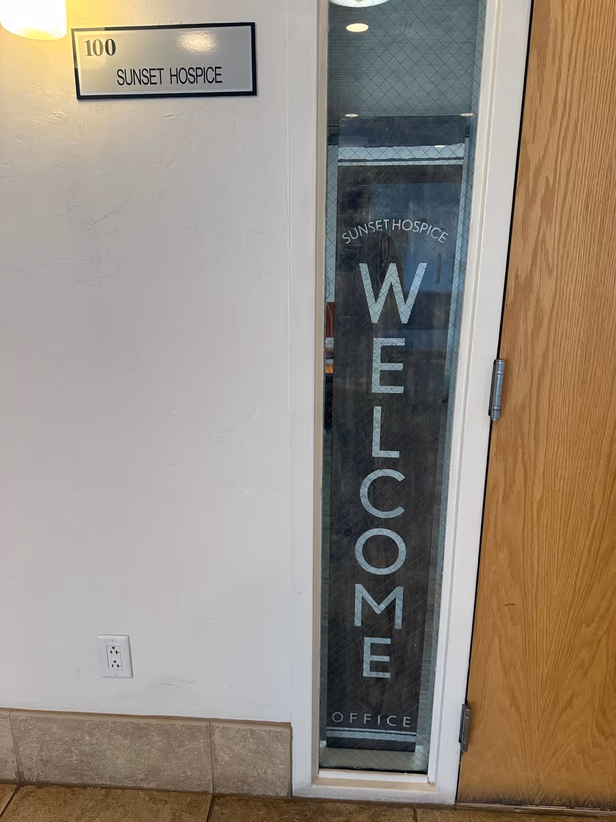 Entrance corridor showing a glass door with a vertical welcome sign and Sunset Hospice plaque.