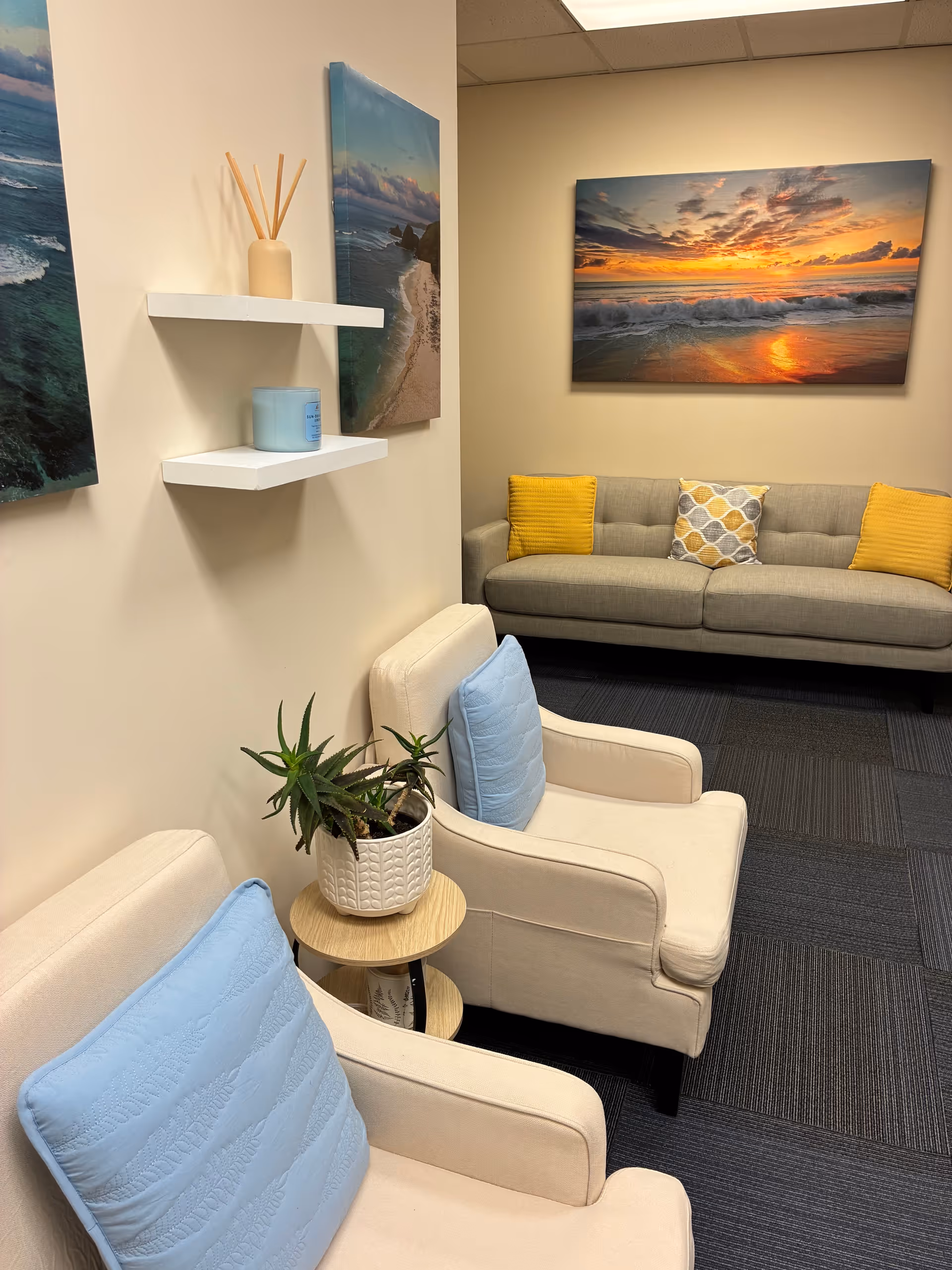 A cozy waiting area with white armchairs, a gray sofa, a plant, and coastal wall art.