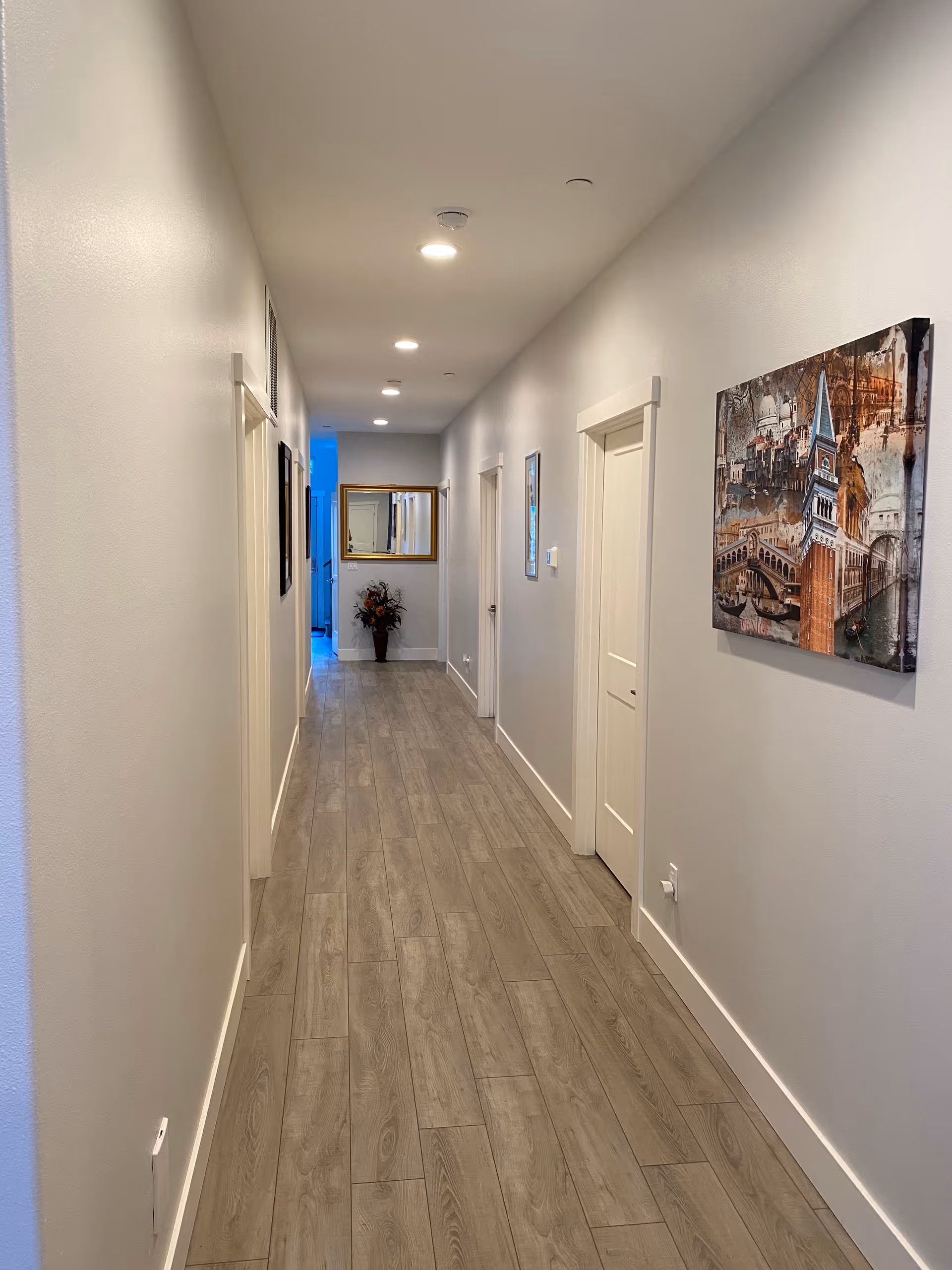Long hallway inside a senior living facility with doors, wall art, and wooden flooring.