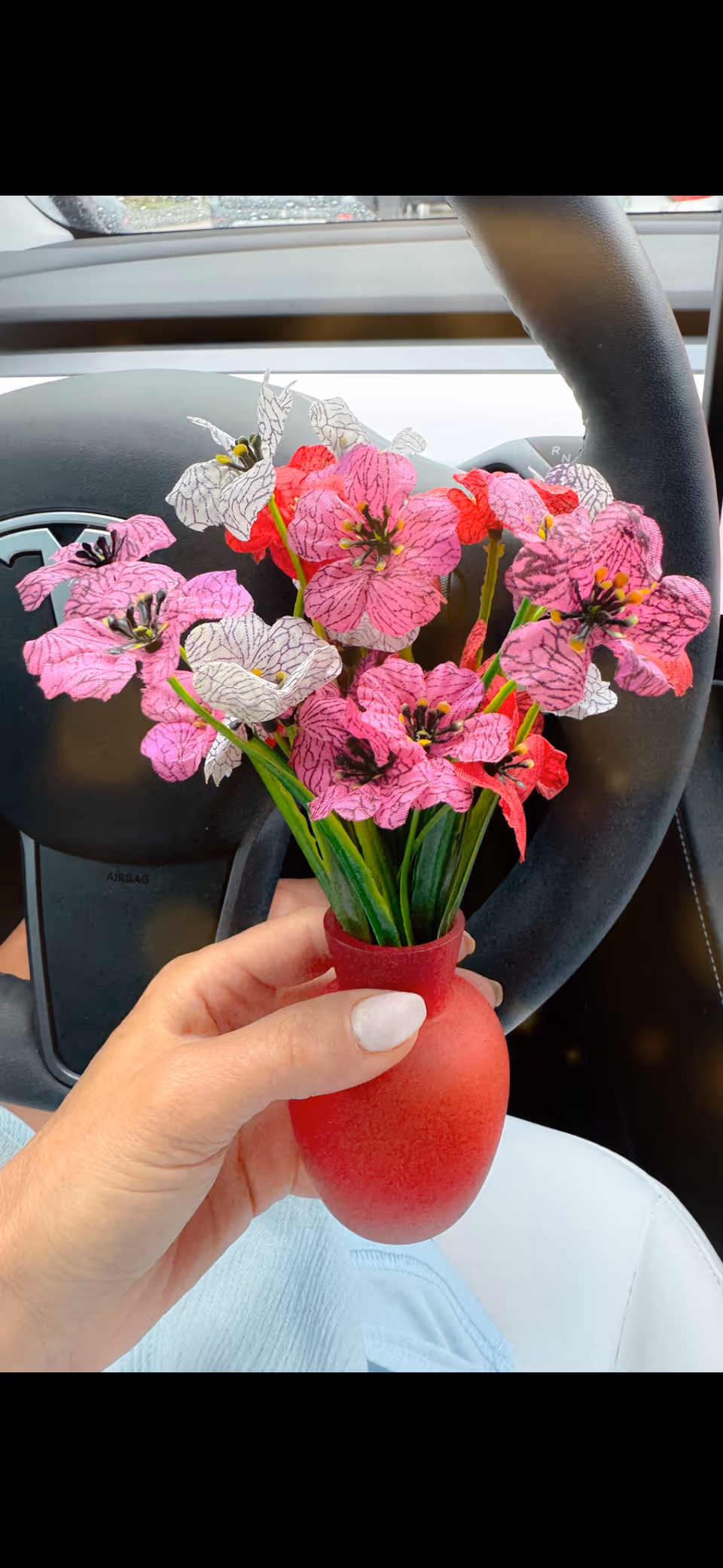 Person's hand holds a red vase with pink flowers inside a car.