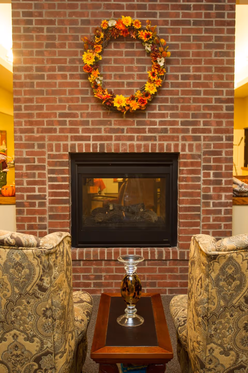 Brick fireplace with autumn wreath flanked by patterned chairs in a cozy living room.