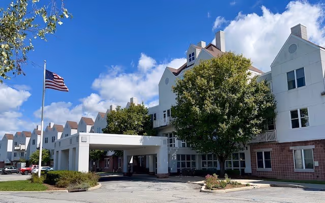 Two-story senior living building with white facade, trees, and an American flag.
